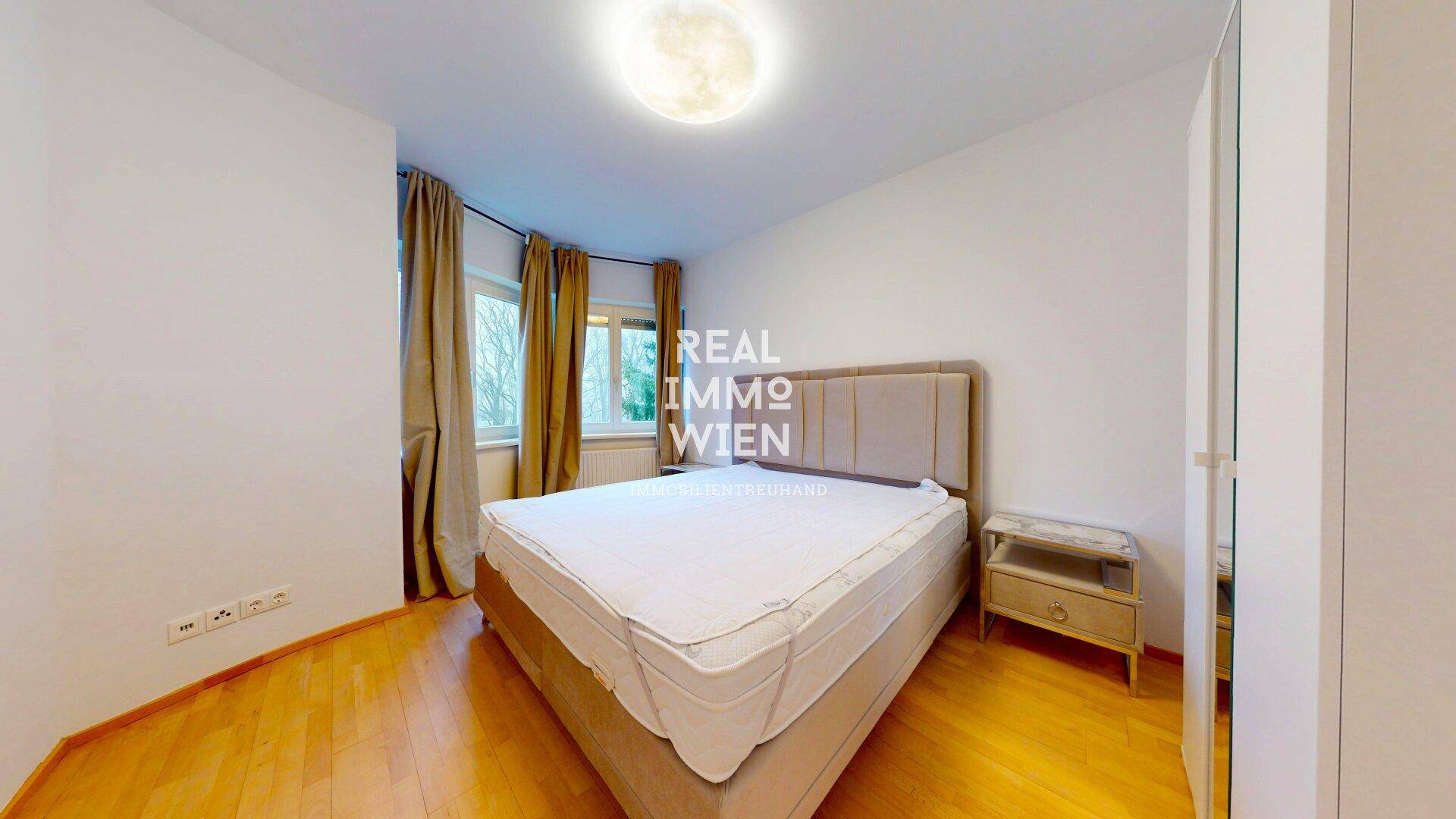Schlafzimmer mit beigem Doppelbett, Holzboden und Fenster für natürliche Belichtung.