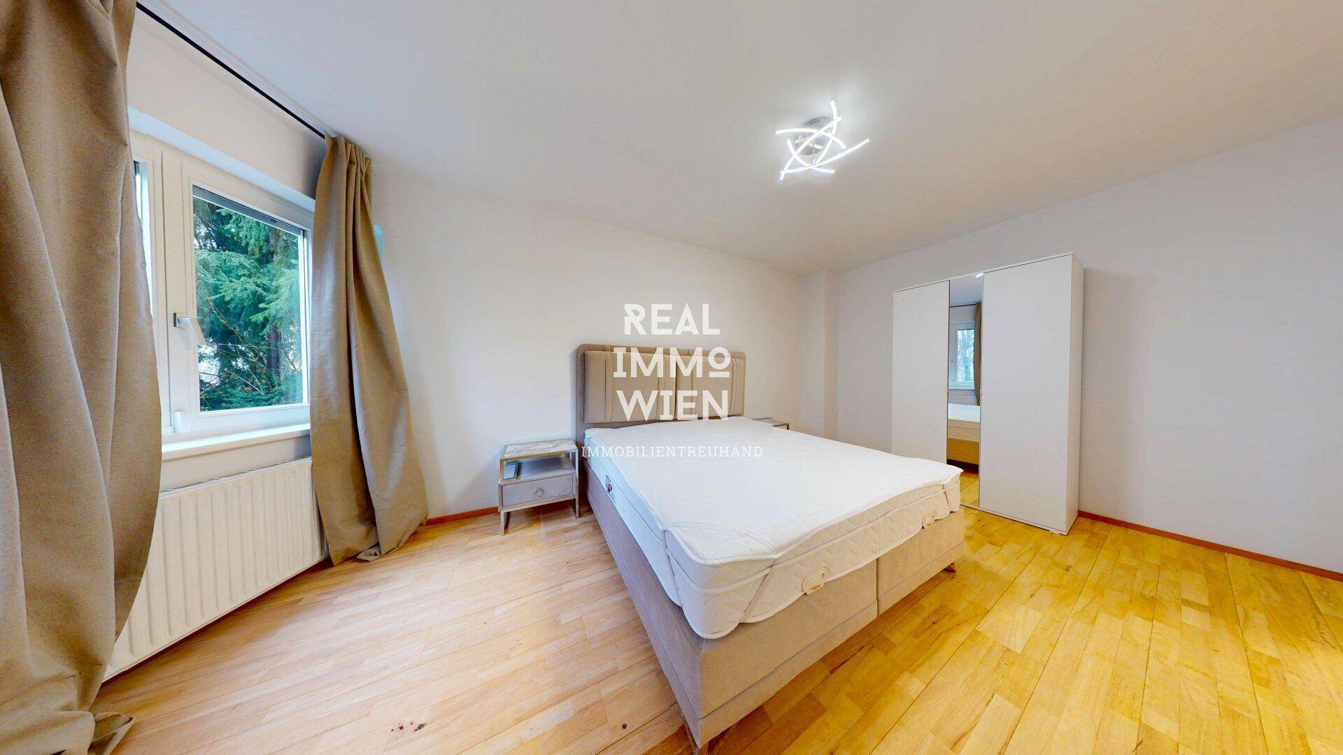 Schlafzimmer mit beigem Doppelbett, Holzboden und Fenster mit Blick nach draußen.