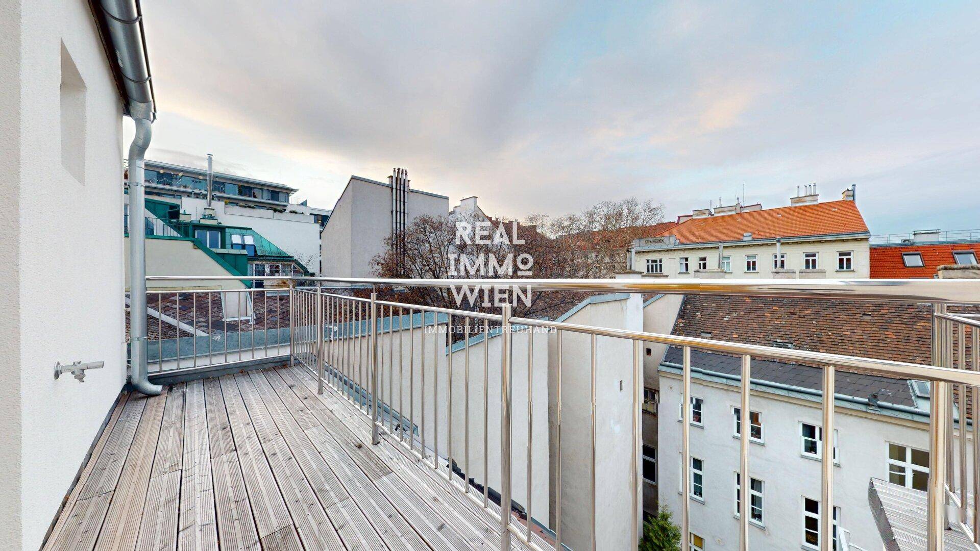 Balkon mit Holzboden und Metallgeländer, bietet einen Blick auf die umliegenden Dächer und die städtische Umgebung.