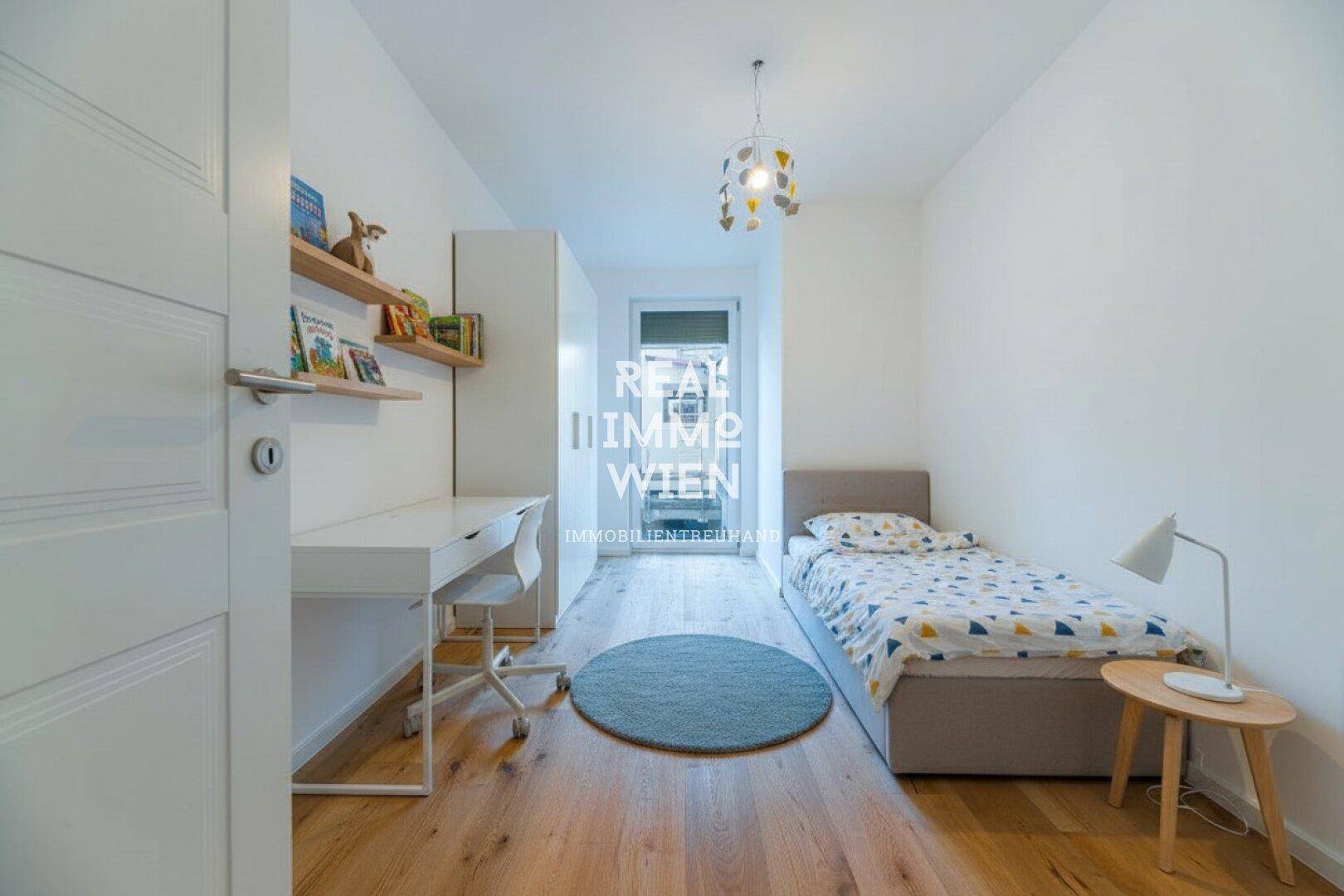 Schmales Kinderzimmer mit Holzboden, Schreibtisch und Einzelbett, gut beleuchtet.