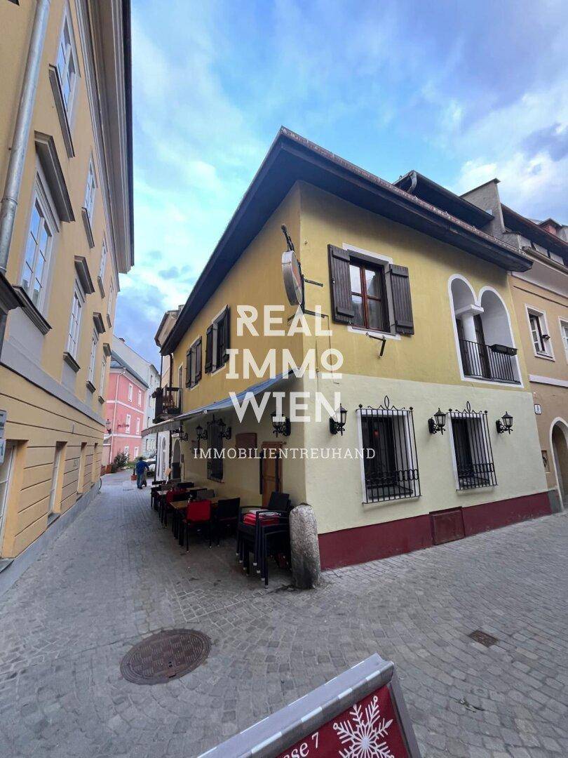Eckgebäude mit gelber Fassade, traditionellen Fensterläden und Tischen im Freien in einer historischen Gasse.