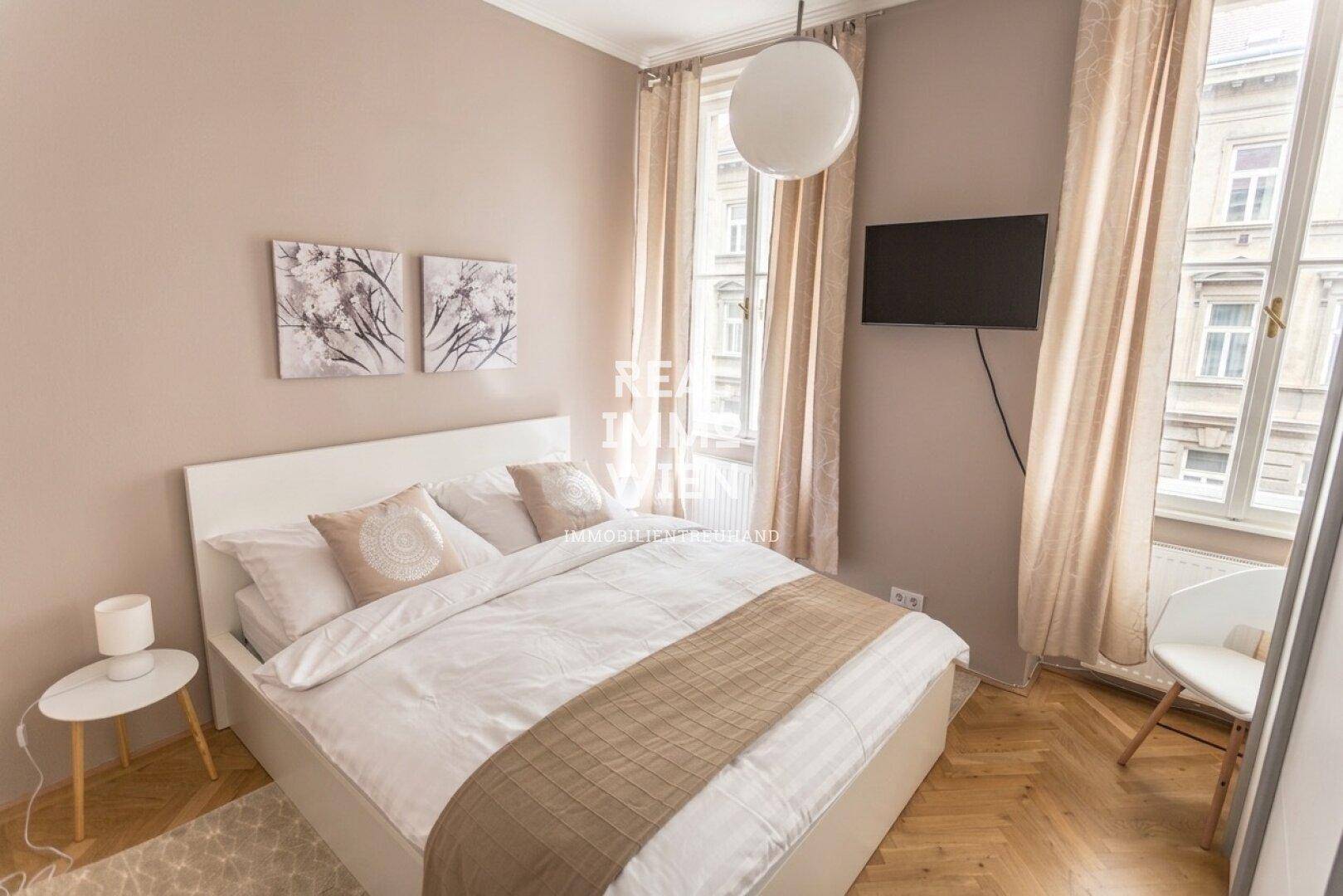 Gemütliches Schlafzimmer mit hellem Bett, Wandfernseher und Vorhängen, die für eine warme Atmosphäre sorgen.