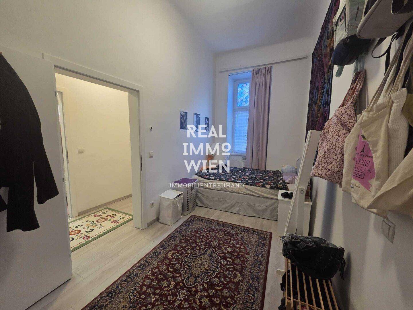 Schlafzimmer mit Bett, Teppich und Zugang zu einem weiteren Raum, mit hellem Laminatboden.
