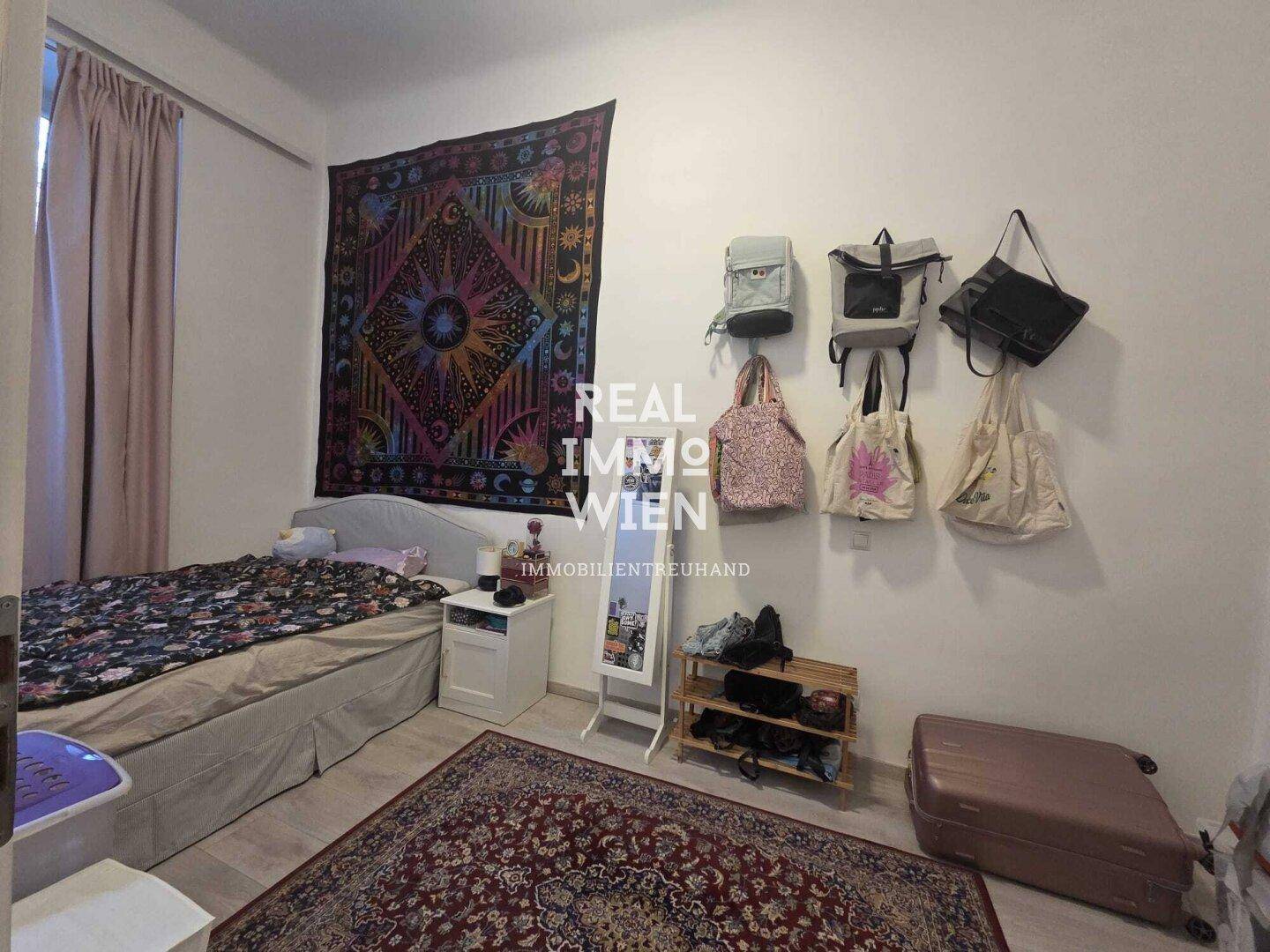 Schlafzimmer mit Bett, Wandteppich und Spiegel, mit hellem Laminatboden und Fenster.