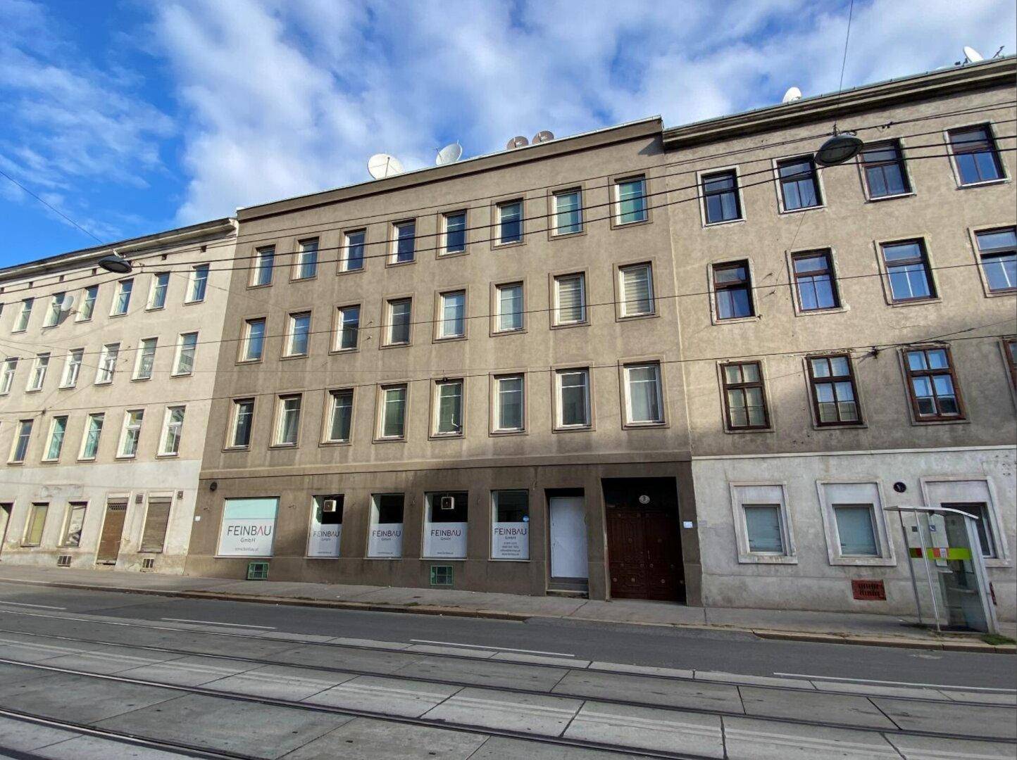 Straßenansicht eines Mehrfamilienhauses mit Geschäften im Erdgeschoss und Straßenbahnschienen.