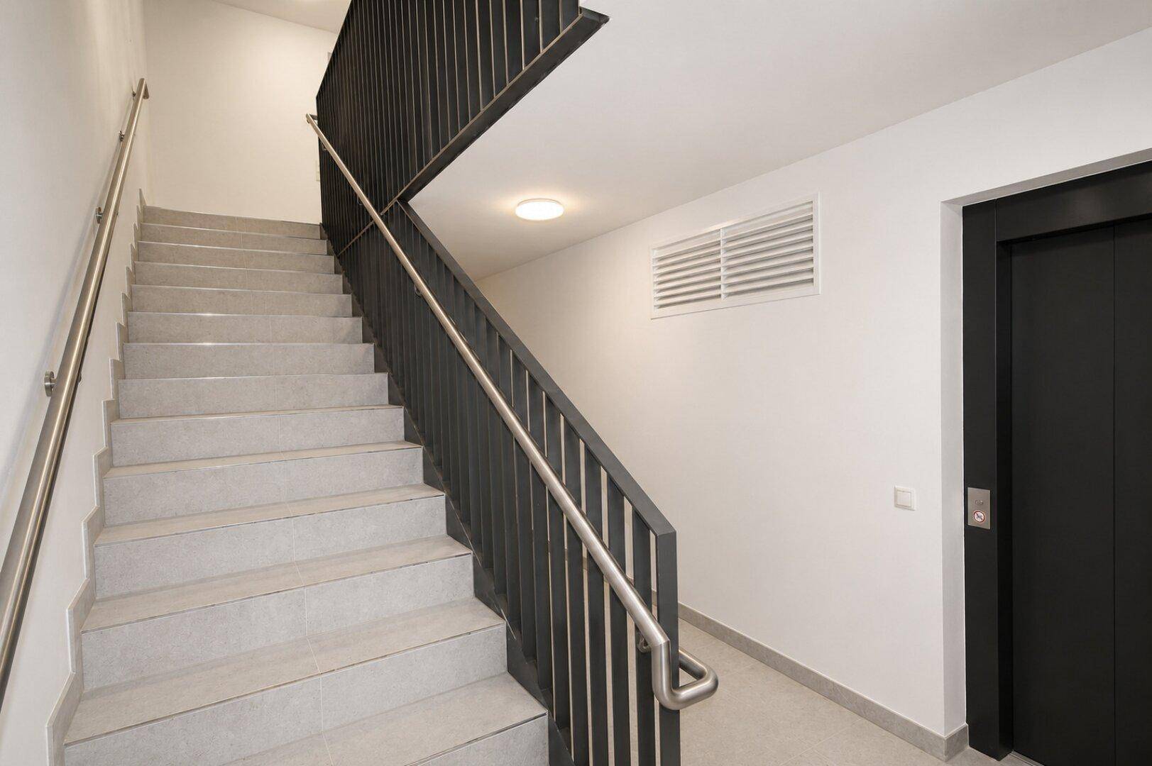 Treppenhaus mit Fliesenstufen, Metallgeländer und einem Aufzug in einem modernen Gebäude.