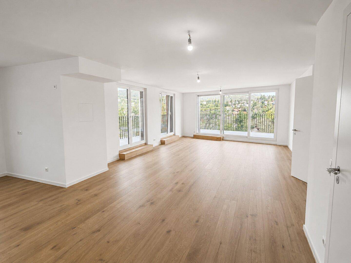 Helles, leeres Wohnzimmer mit Holzboden und großen Fenstern, die viel Tageslicht hereinlassen.