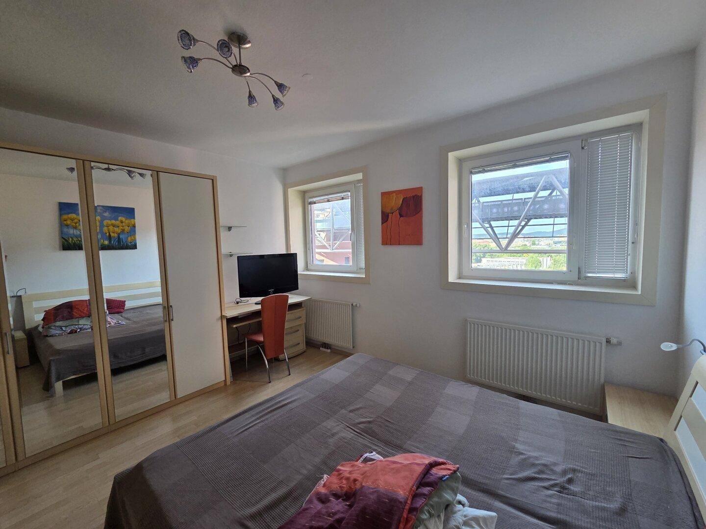 Schlafzimmer mit Doppelbett, Kleiderschrank mit Spiegeltüren und zwei Fenstern für Tageslicht.