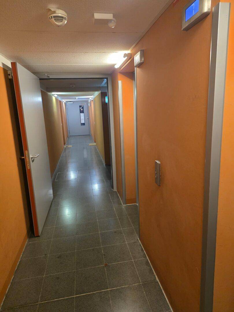 Langer Flur in einem Mehrfamilienhaus mit orangefarbenen Wänden und Fliesenboden.
