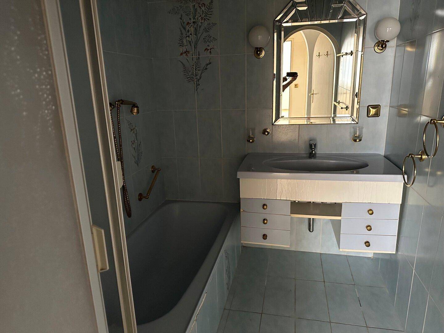 Badezimmer im Stil der 80er Jahre mit hellblauen Fliesen, Badewanne und Waschtisch mit Spiegel.