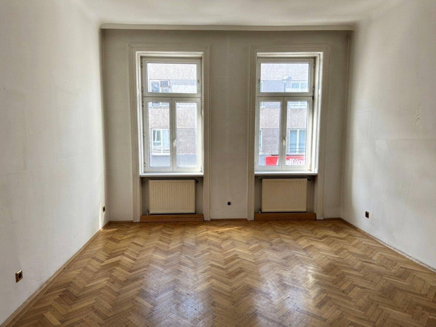 Helles Zimmer mit Fischgrätparkett und zwei hohen Fenstern mit Blick auf die Stadt.