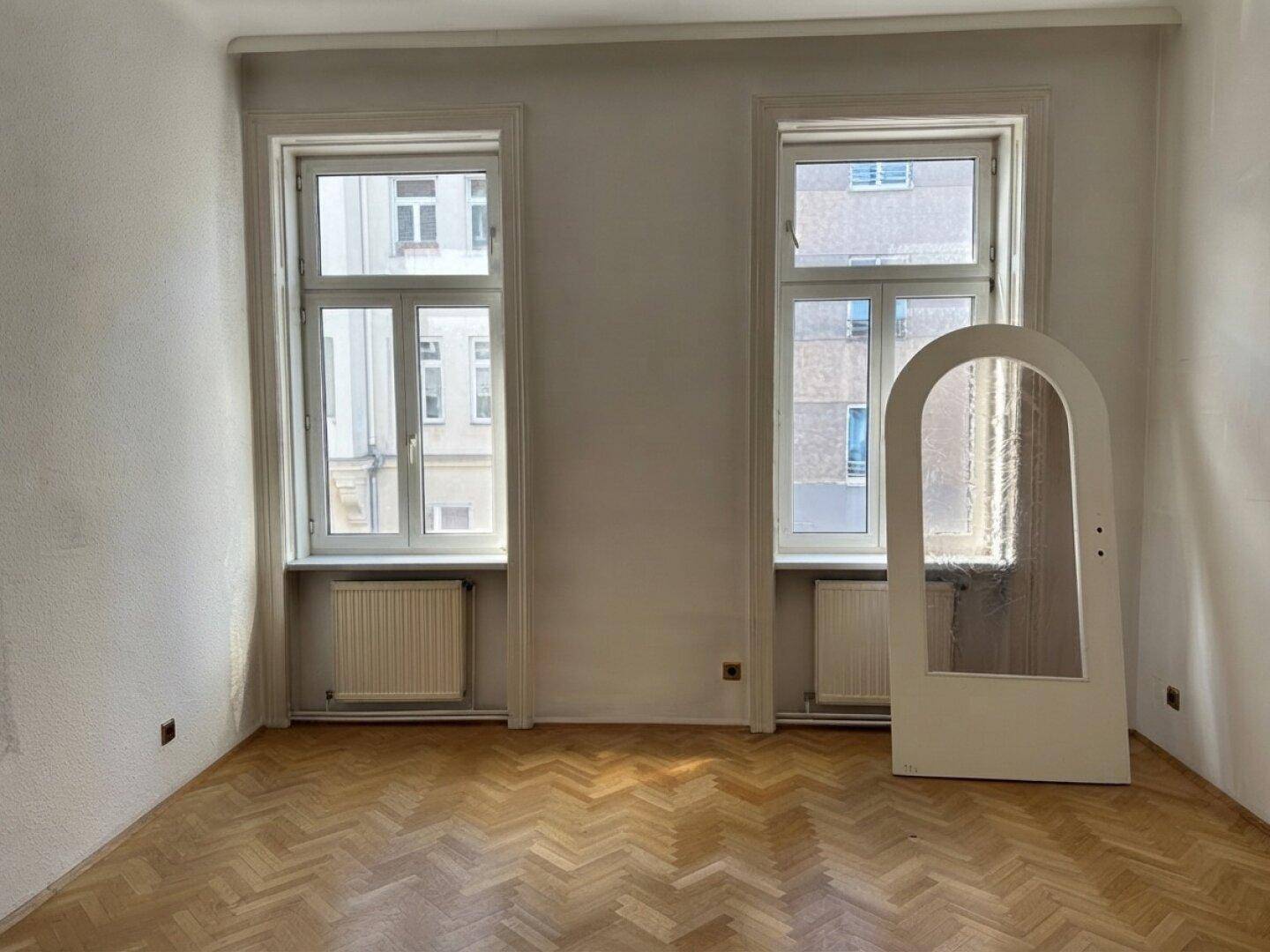 Leeres Zimmer mit Fischgrätparkett, hohen Fenstern und einer dekorativen Bogentür.