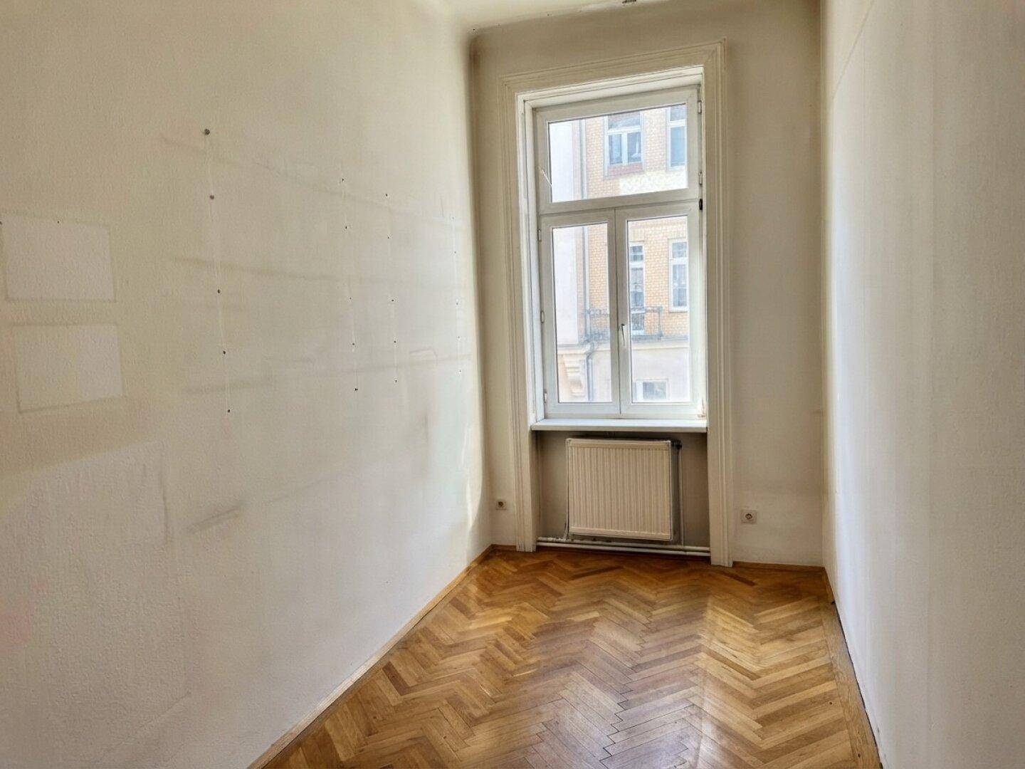 Schmaler Raum mit Fischgrätparkett und einem hohen Fenster, das viel Tageslicht hereinlässt.