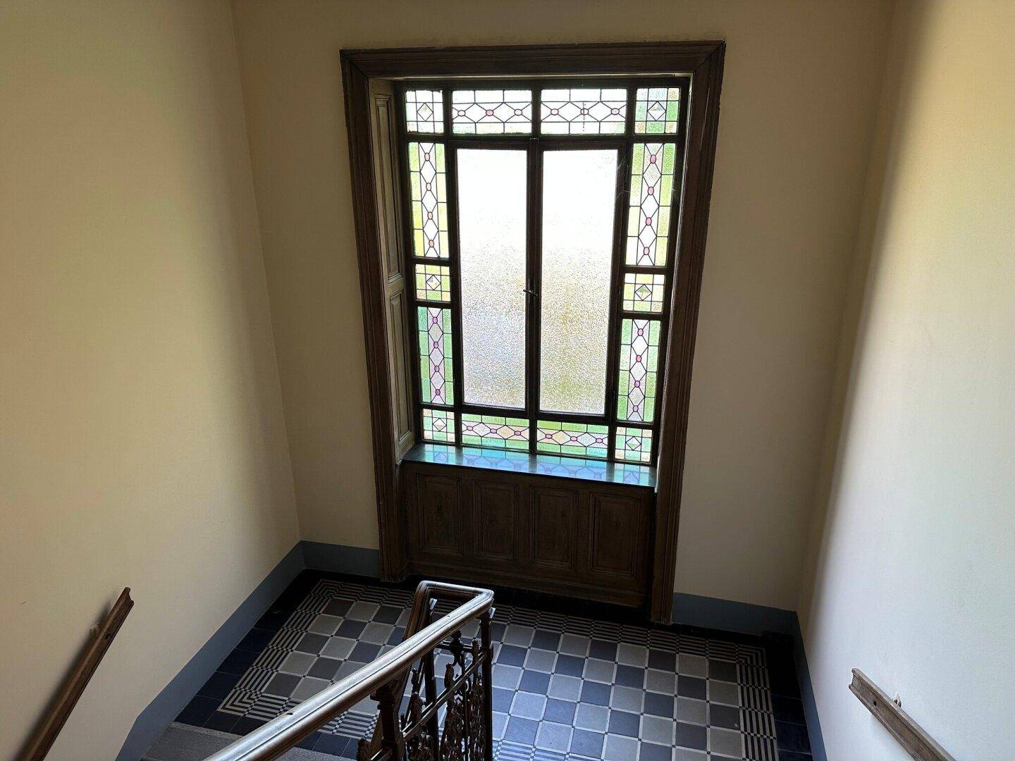 Historisches Treppenhaus mit Buntglasfenster, gemusterten Fliesen und schmiedeeisernem Geländer.