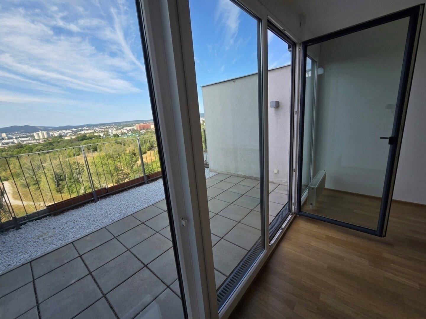 Innenansicht eines Raumes mit Holzboden und großen Glastüren, die zur Terrasse mit Weitblick führen.