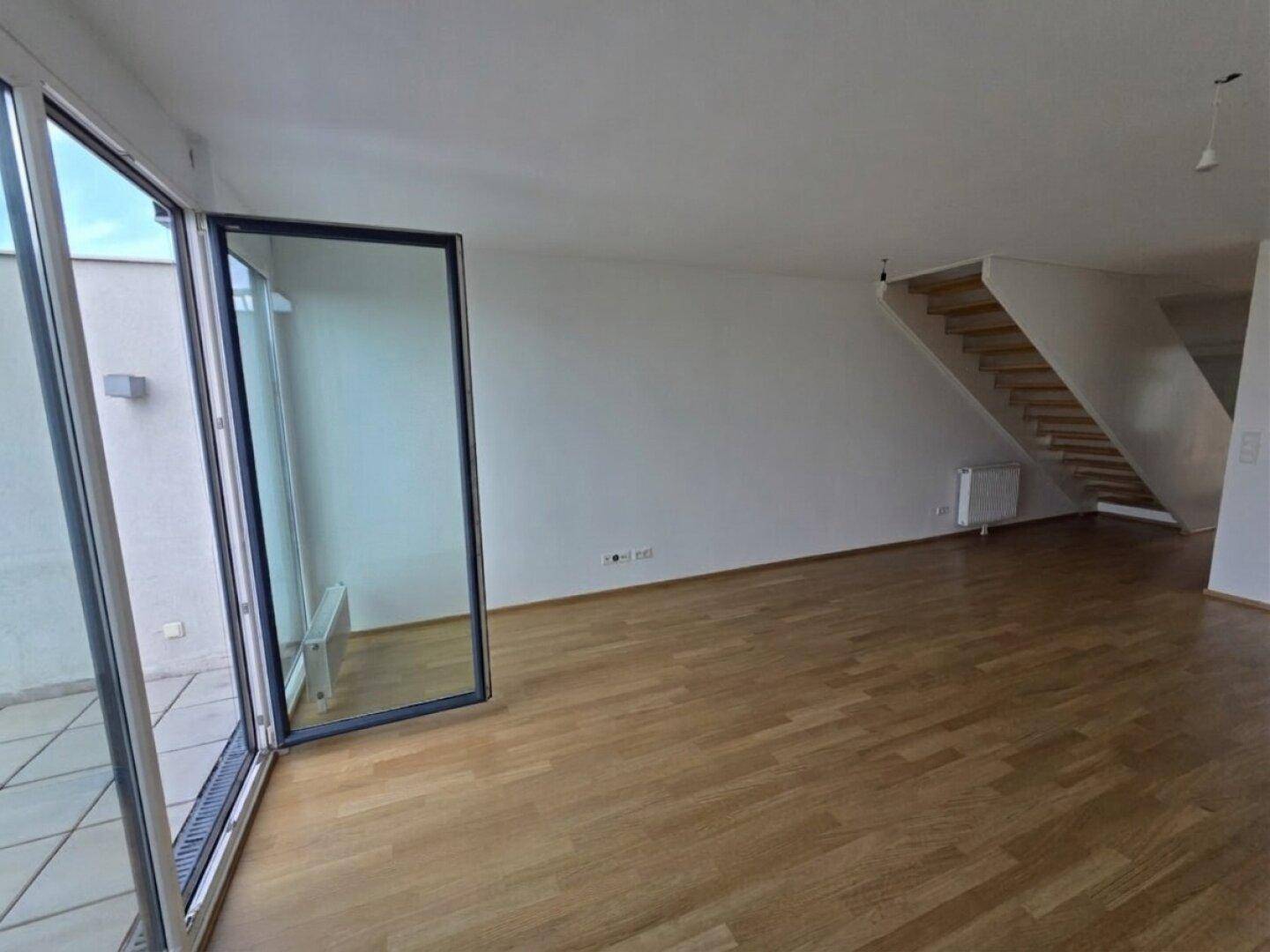 Heller Wohnbereich mit Parkettboden, Zugang zur Terrasse und einer modernen Treppe im Hintergrund.