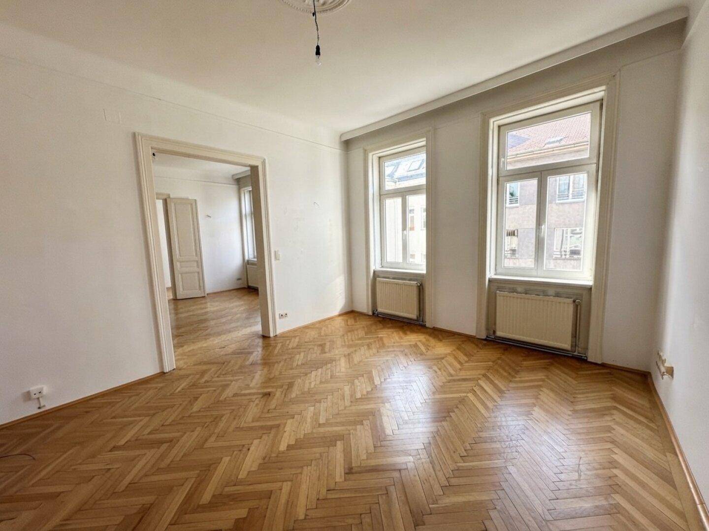 Geräumiges Zimmer mit Fischgrätparkett und hohen Fenstern, das viel Tageslicht hereinlässt.
