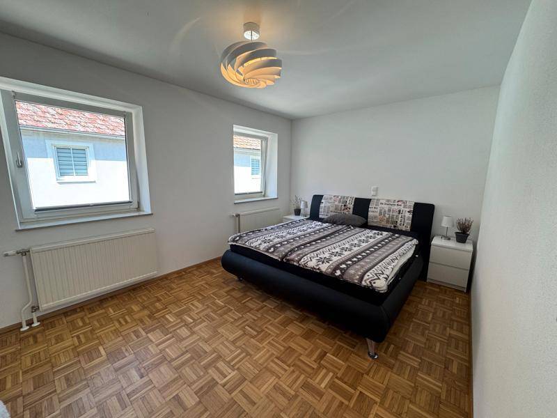 Helles Schlafzimmer mit Doppelbett, Parkettboden und zwei Fenstern für viel Tageslicht.