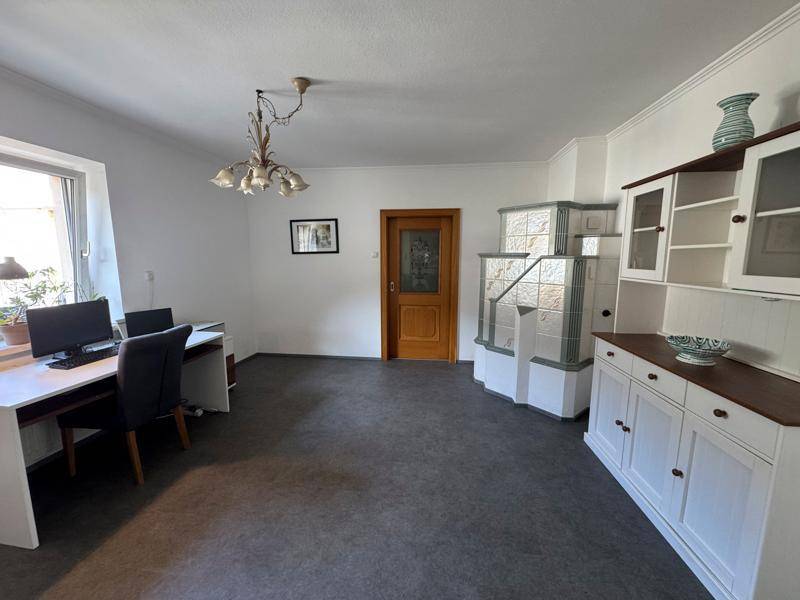 Geräumiges Wohnzimmer mit Arbeitsbereich, traditionellem Kachelofen und Vinylboden.
