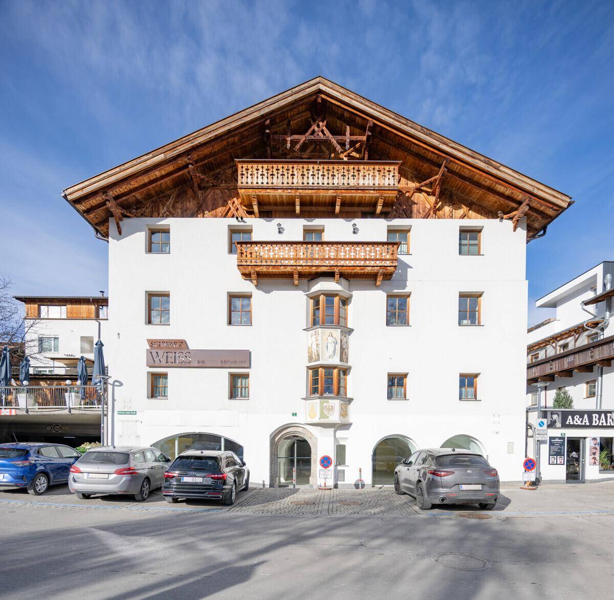 Traditionelles Gebäude mit Holzbalkonen, weißer Fassade und Giebeldach in sonniger Lage mit Parkplätzen.