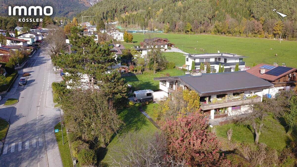 Luftaufnahme eines Wohnhauses mit Flachdach, Balkon, angrenzender Straße und weitem Blick ins Grüne und auf die Berge.