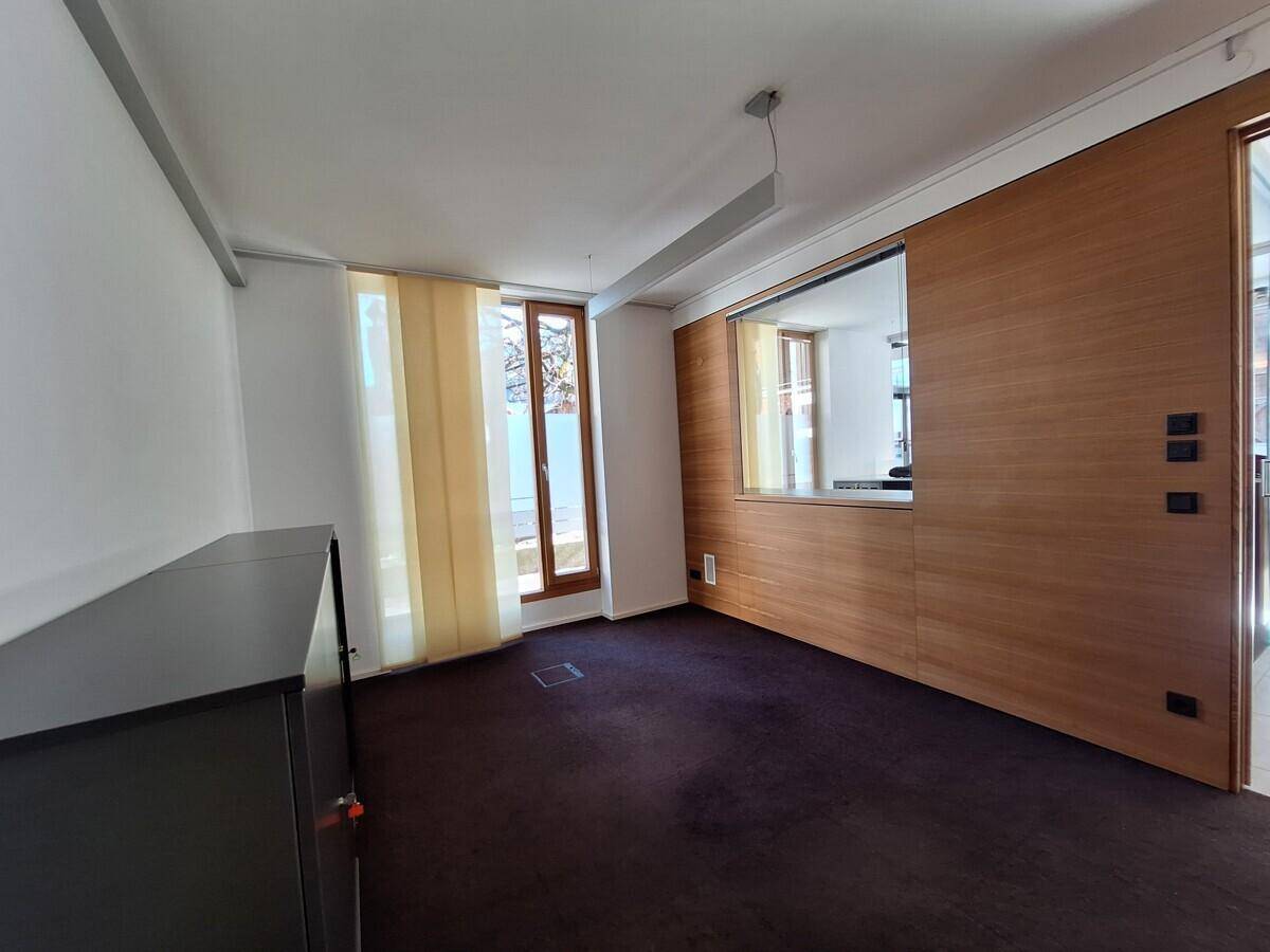 Büroraum mit dunklem Teppichboden, weißen Wänden und einer Holzwand mit integriertem Fenster.
