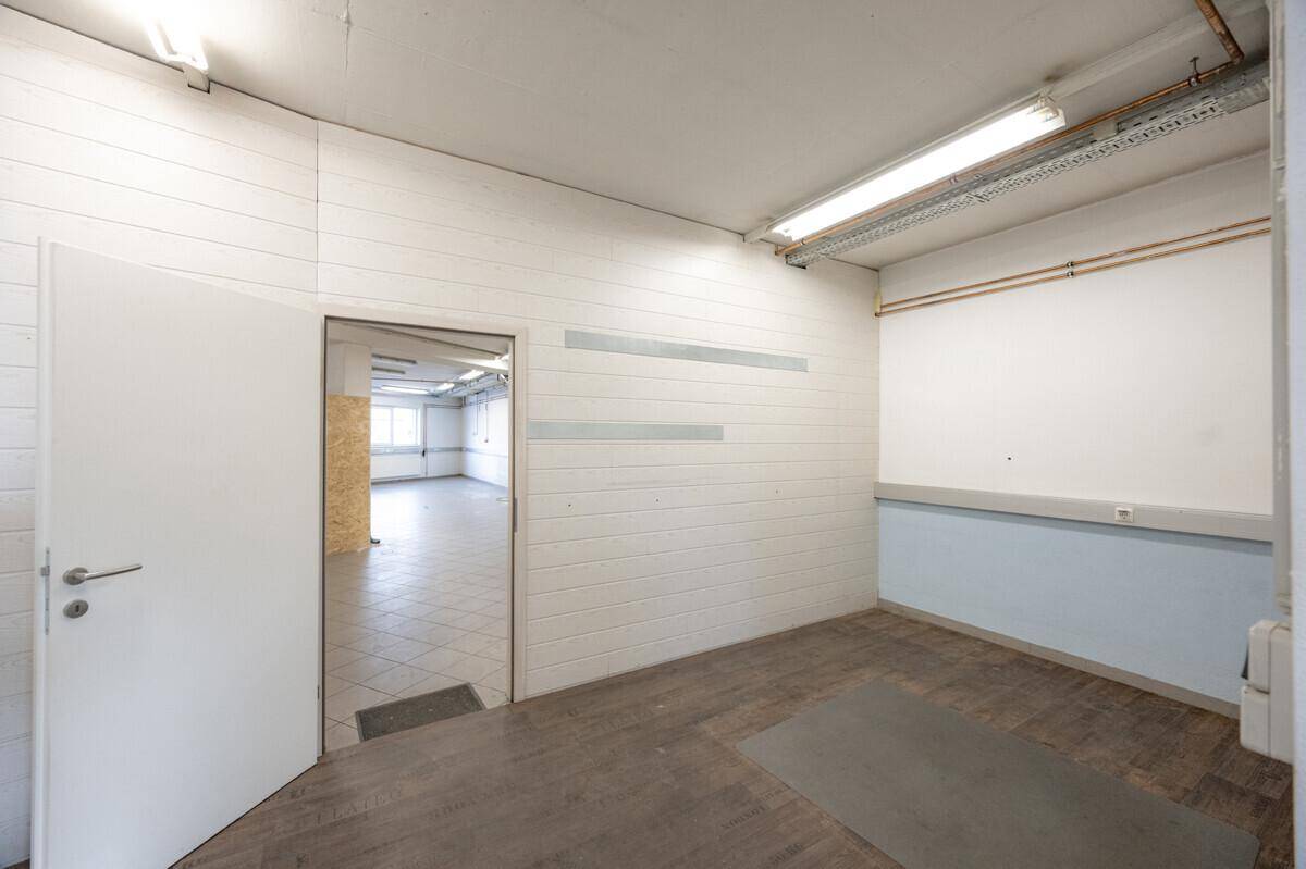 Kleinerer Raum mit Holzwänden, einer Tür zu einem größeren Bereich und einem dunklen Bodenbelag.