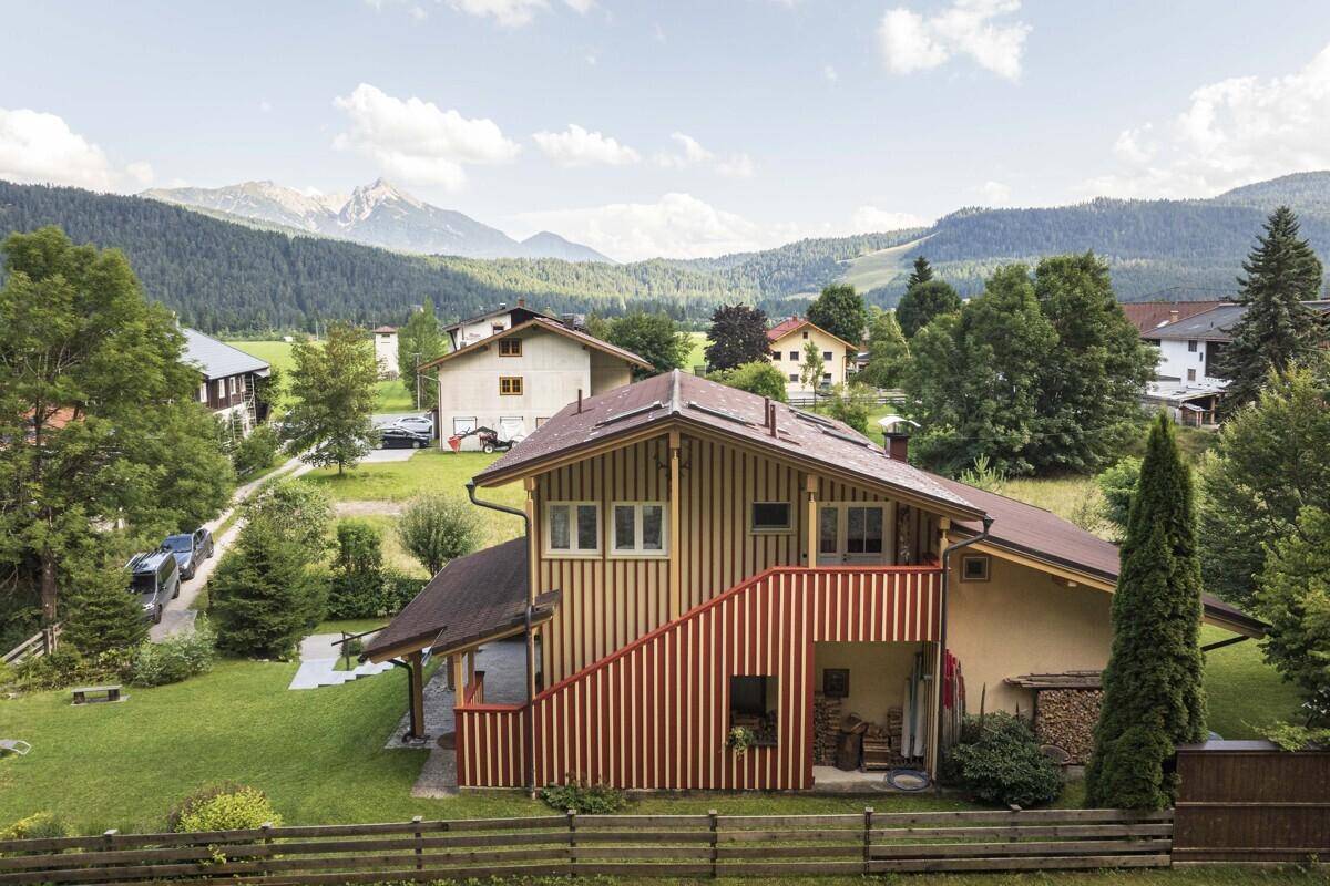 Traditionelles Haus mit Holzfassade und roter Treppe, umgeben von grüner Natur und Bergen.
