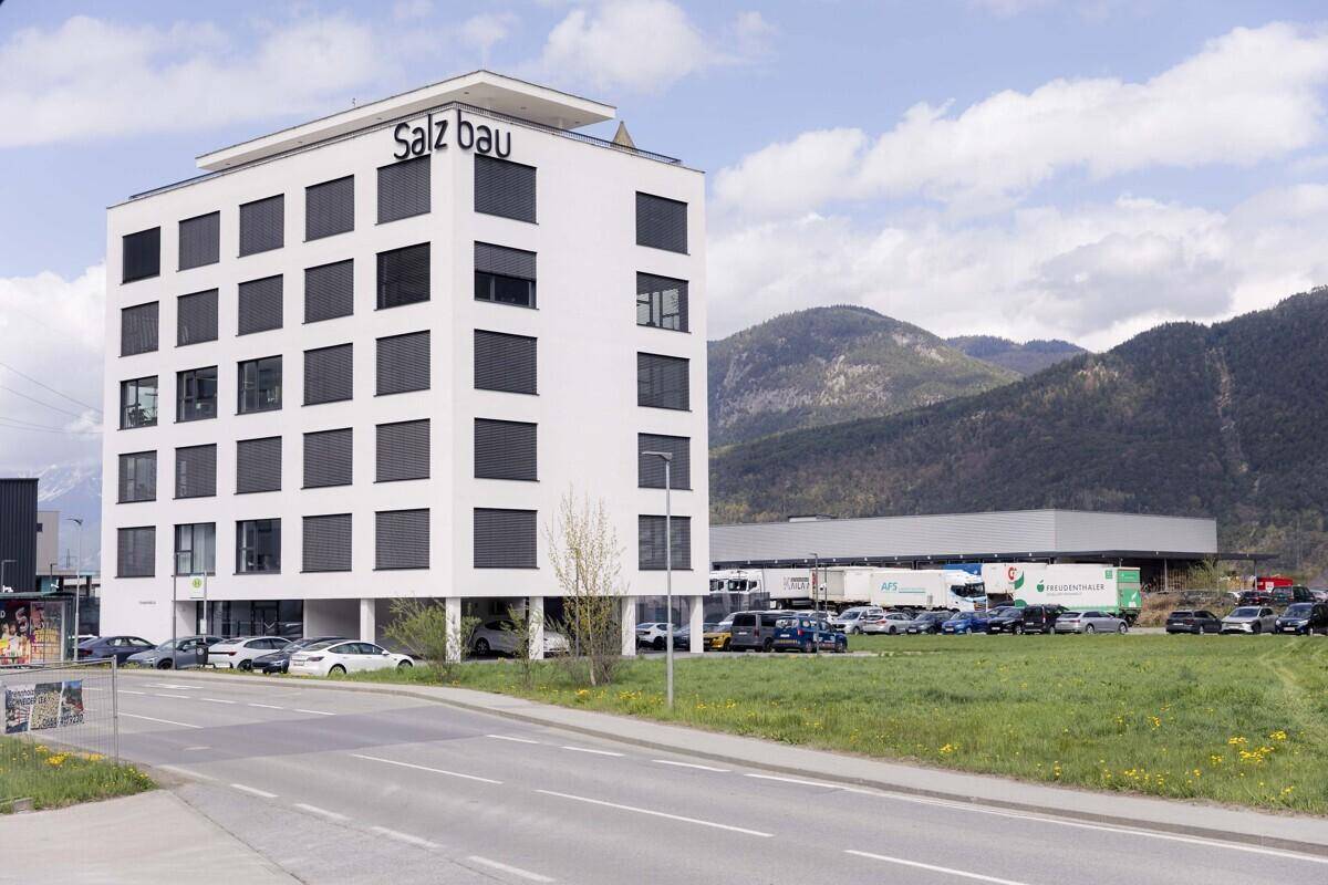 Modernes weißes Bürogebäude mit großen Fenstern und Parkplätzen vor einer Bergkulisse.