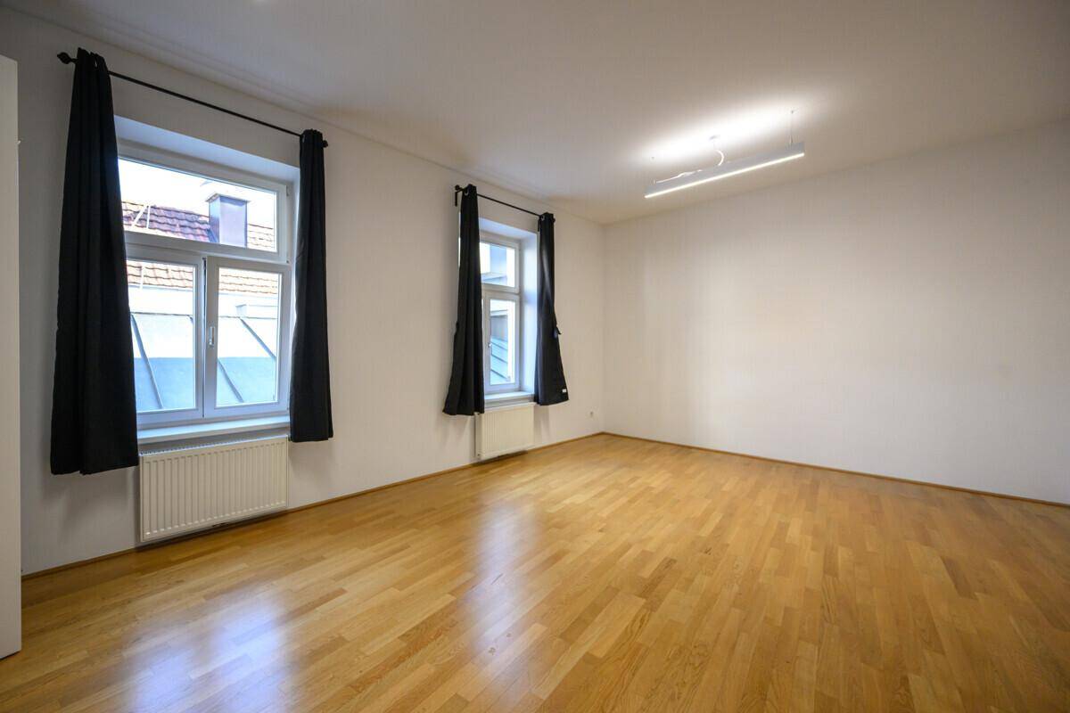 Geräumiges, leeres Zimmer mit zwei großen Fenstern und dunklen Vorhängen, ideal als Wohnzimmer.