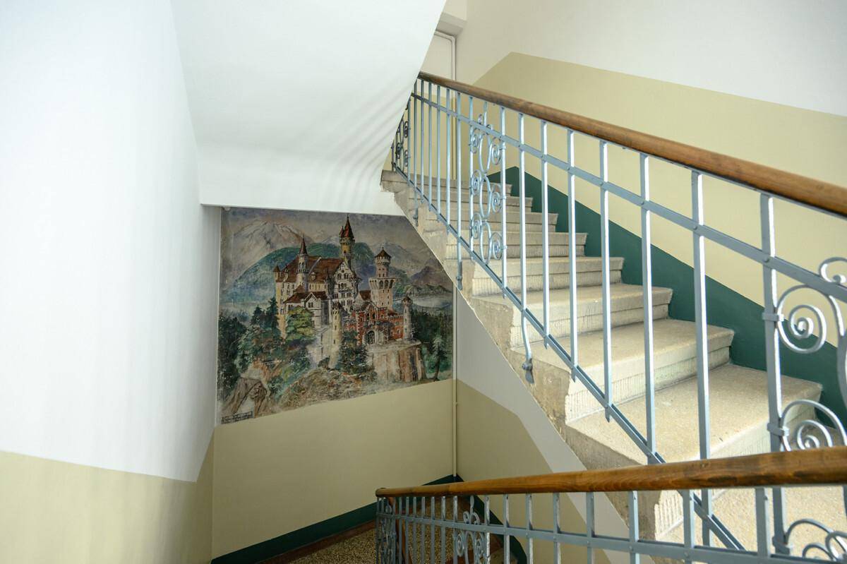 Treppenhaus mit Steinstufen, schmiedeeisernem Geländer und Wandgemälde eines Schlosses.