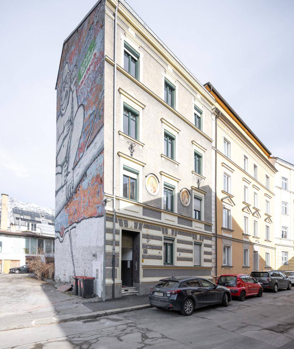 Historisches Gebäude mit markanter Fassade, Wandgemälde und parkenden Autos auf der Straße.