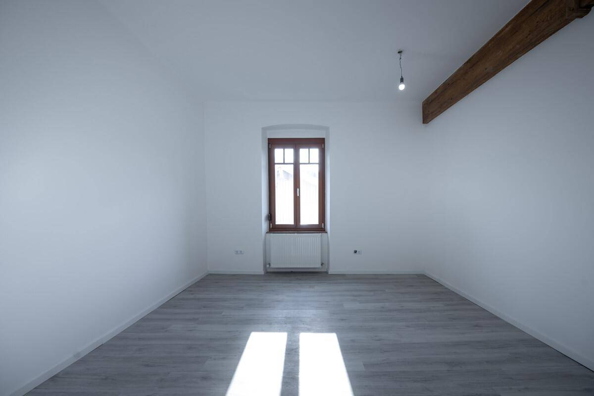 Leerer Raum mit hellem Laminatboden, einem Fenster und einem Heizkörper.