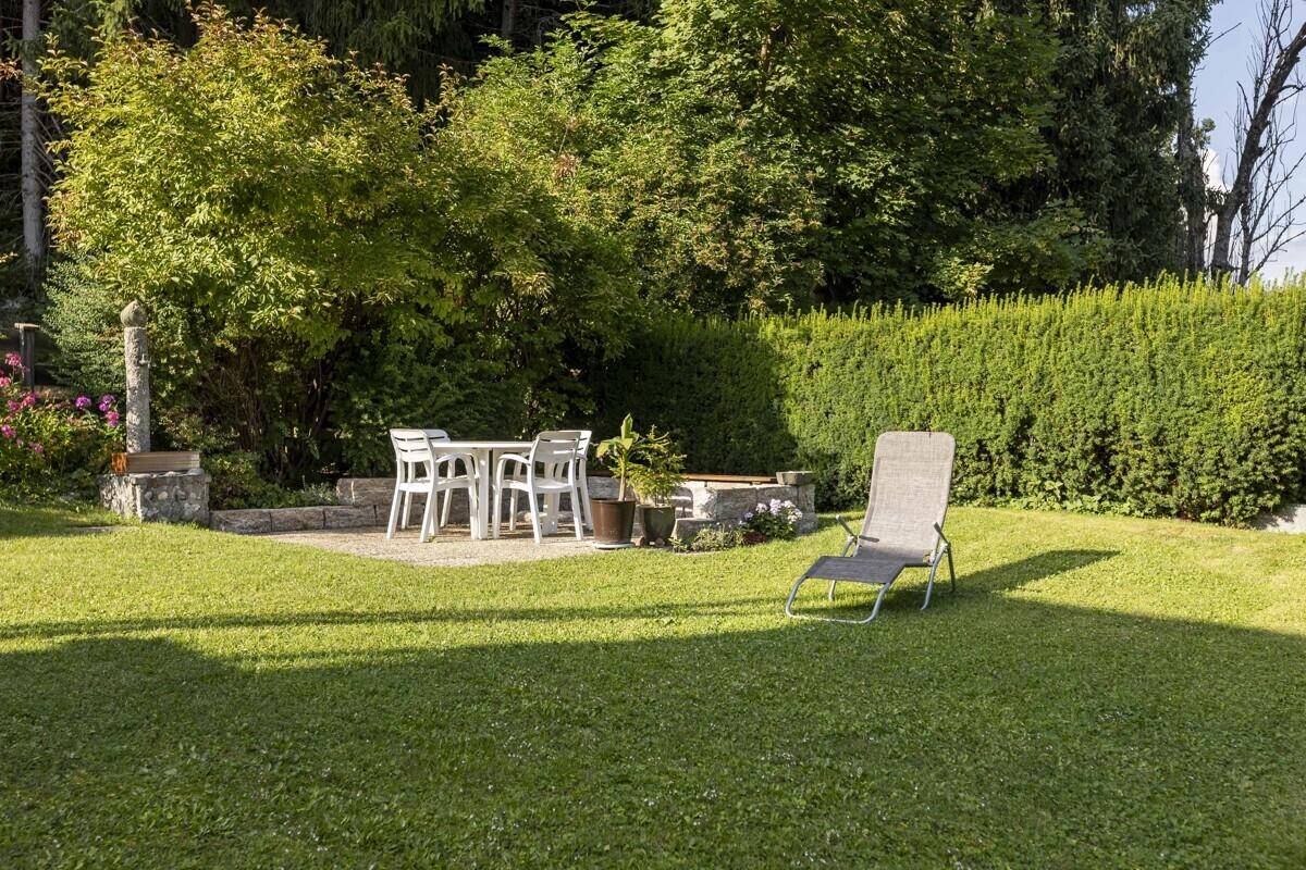 Gemütlicher Sitzbereich im Garten mit Steintisch und Liegestuhl, umgeben von Hecken.
