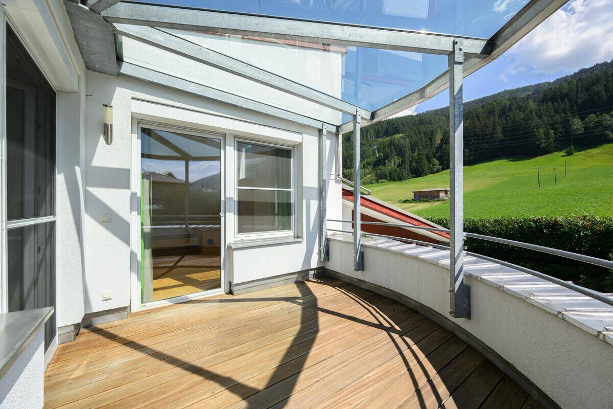 Großer, geschwungener Balkon mit Holzbelag, Glasdach und Blick ins Grüne.