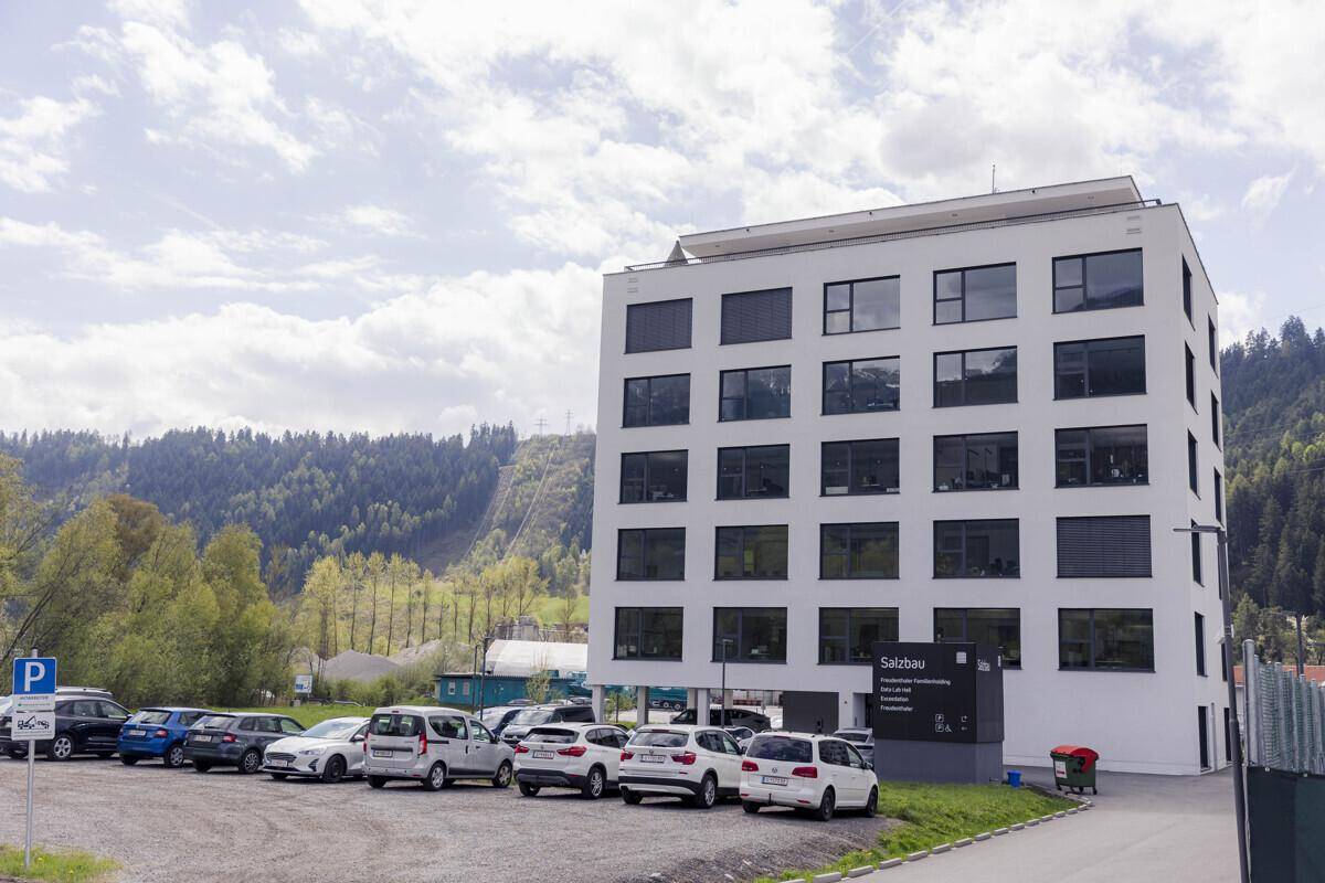 Modernes Bürogebäude in weißer Fassade mit Parkplätzen und grüner Umgebung vor Bergkulisse.