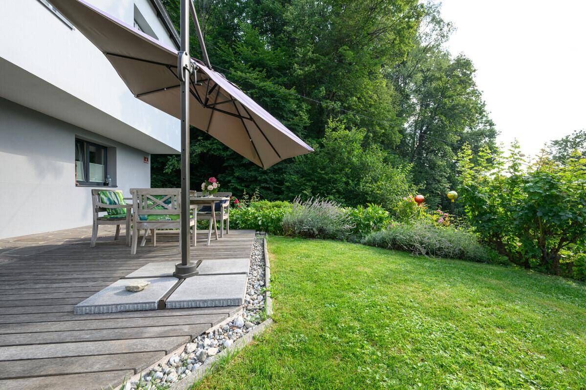 Gepflegter Gartenbereich mit Rasenfläche, Holzterrasse und Sitzgelegenheiten unter einem Sonnenschirm.