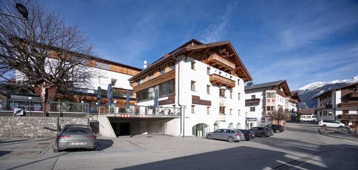 Historisches Gebäude mit Holzbalkonen und traditioneller Architektur in einer Berglandschaft mit Parkplätzen.