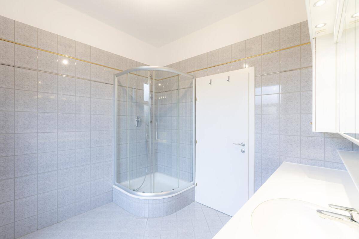 Badezimmer mit Duschkabine, hellgrauen Fliesen und einem Fenster für Tageslicht.