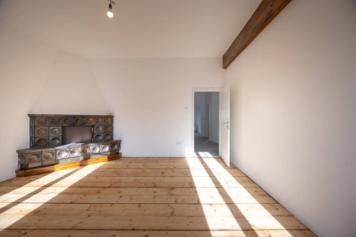 Heller Raum mit Holzdielenboden, sichtbarem Deckenbalken und traditionellem Kachelofen.