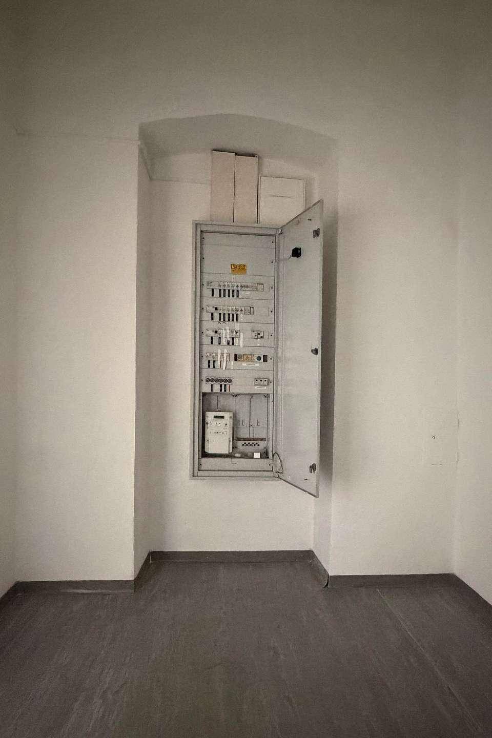 Kleiner Raum mit offenem Sicherungskasten, weißen Wänden und dunklem Vinylboden.