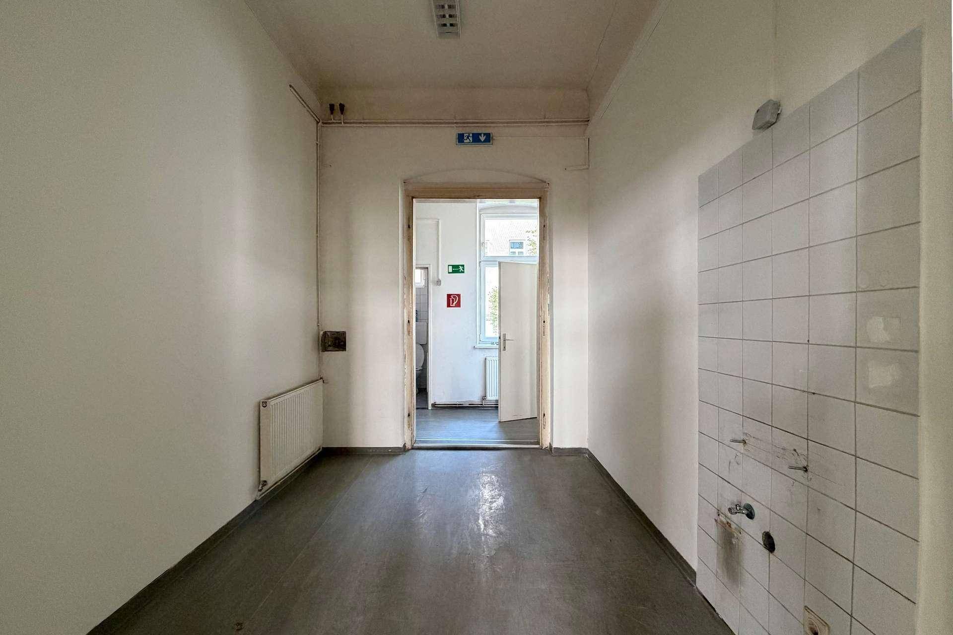 Langer Flur mit dunklem Boden, weißen Wänden und einer Tür zu einem weiteren Raum.
