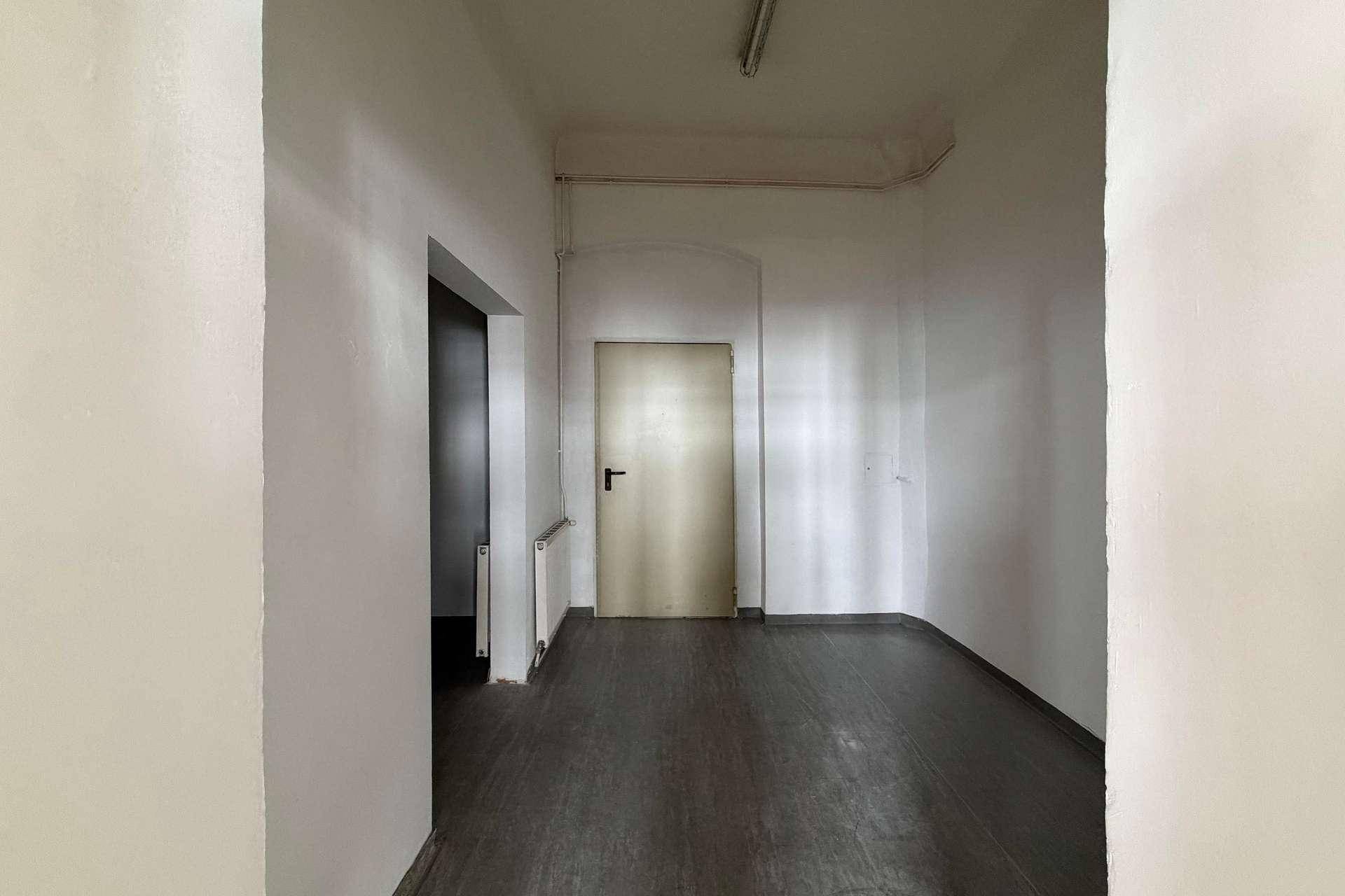 Schmaler Flur mit dunklem Bodenbelag, weißen Wänden und einer einfachen Tür am Ende.