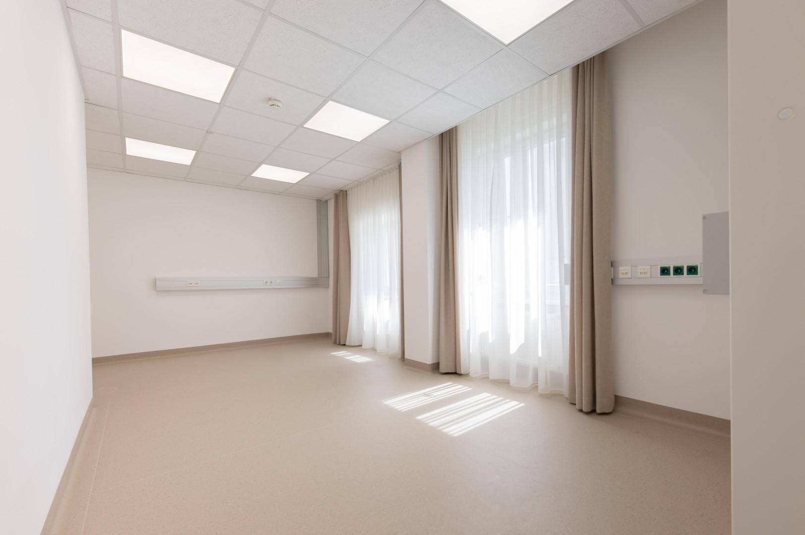 Geräumiger Behandlungsraum mit großen Fenstern, hellen Vorhängen und funktionalen Wandanschlüssen.