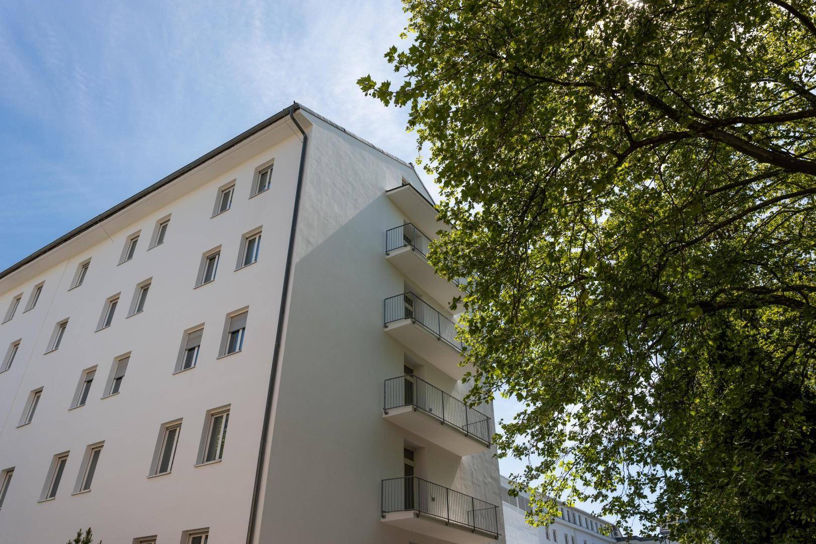 Weiße Gebäudefassade mit vielen Fenstern und kleinen Balkonen, umgeben von grünen Bäumen.