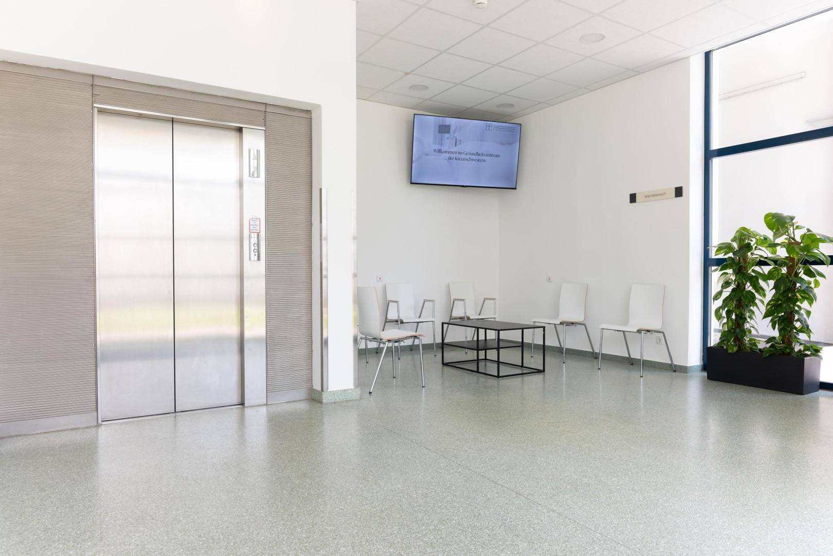 Helle Lobby mit modernem Aufzug, Sitzgelegenheiten und einem Informationsbildschirm an der Wand.