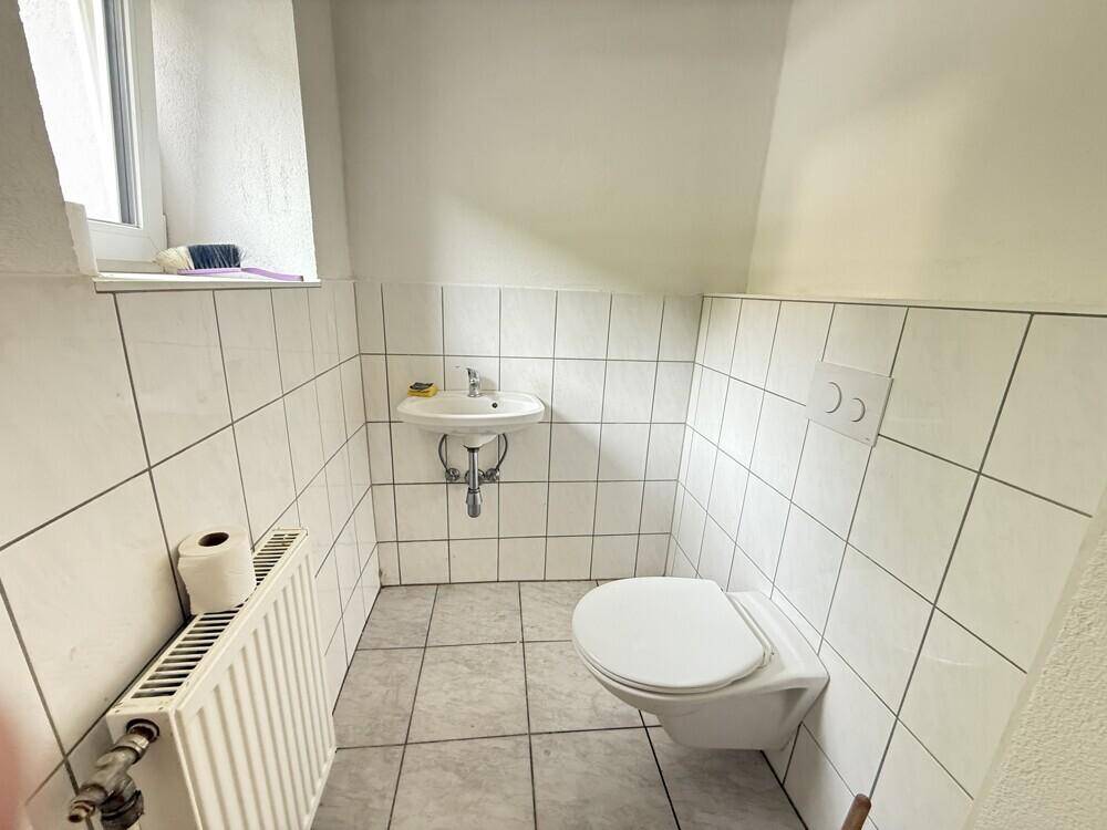 Kleines Gäste-WC mit Fenster, Wand-WC, Waschbecken und weißen Fliesen an Wänden und Boden.