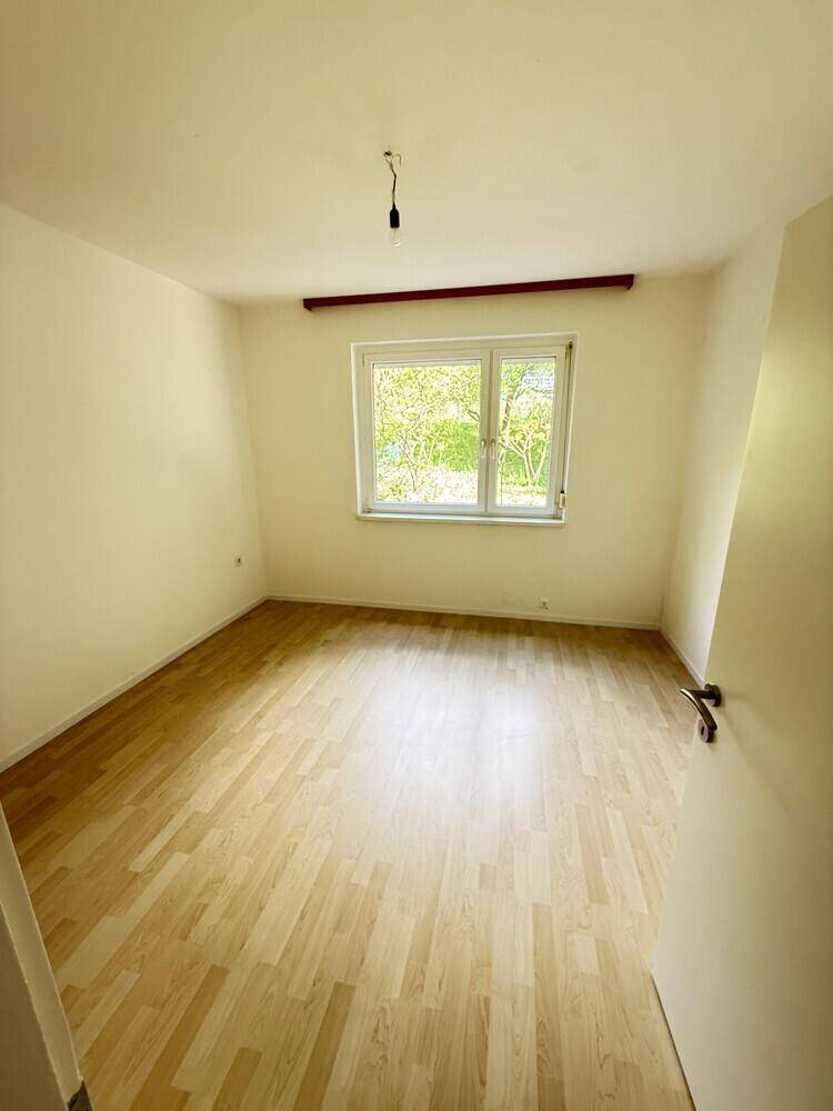 Leeres Schlafzimmer mit hellem Laminatboden, weißen Wänden und einem Fenster mit Blick ins Grüne.