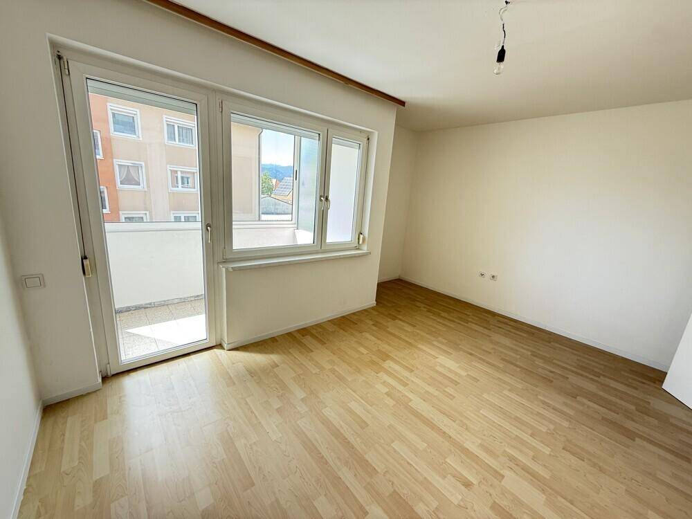 Helles Zimmer mit hellem Laminatboden, weißen Wänden und Zugang zu einem kleinen Balkon.