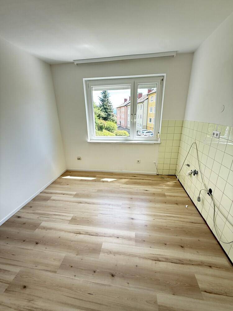 Leerer Küchenbereich mit hellem Laminatboden, weißen Wänden und gelben Fliesen an einer Wand.
